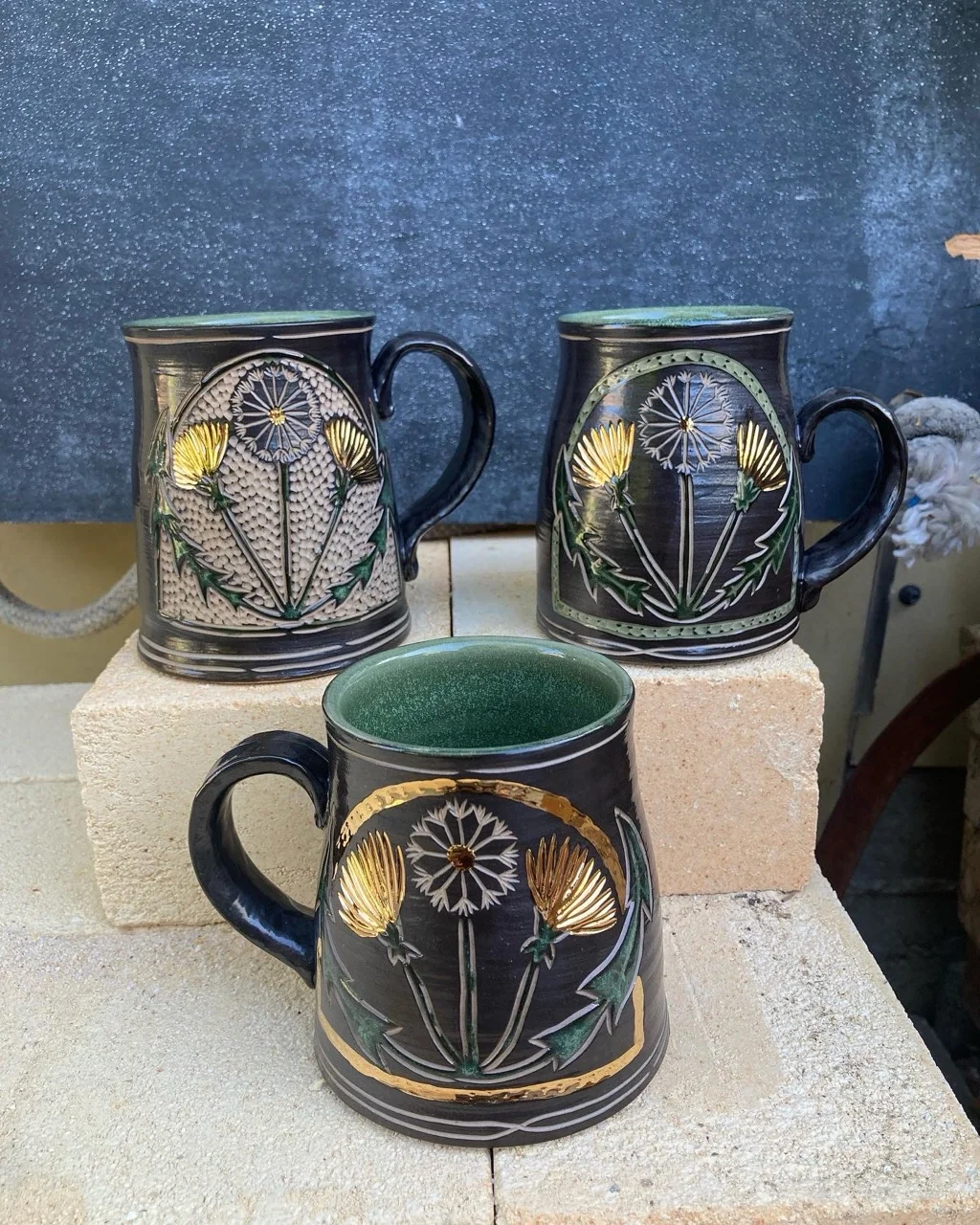 Three black mugs with green interiors, with golden dandelions carved and painted